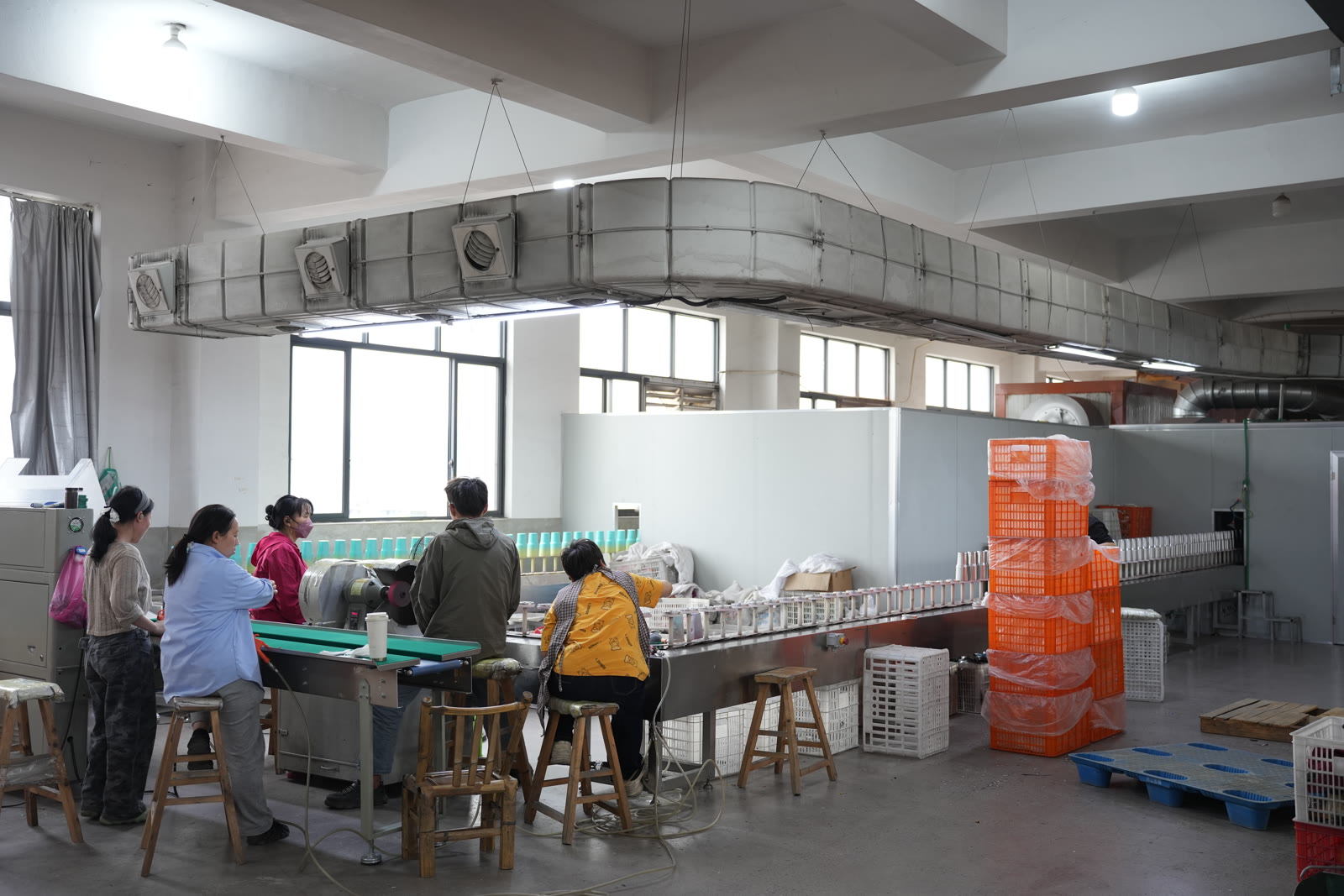 Workers seated along an assembly conveyor line in a tumbler factory, with orange crates of components stacked at the end of the line
